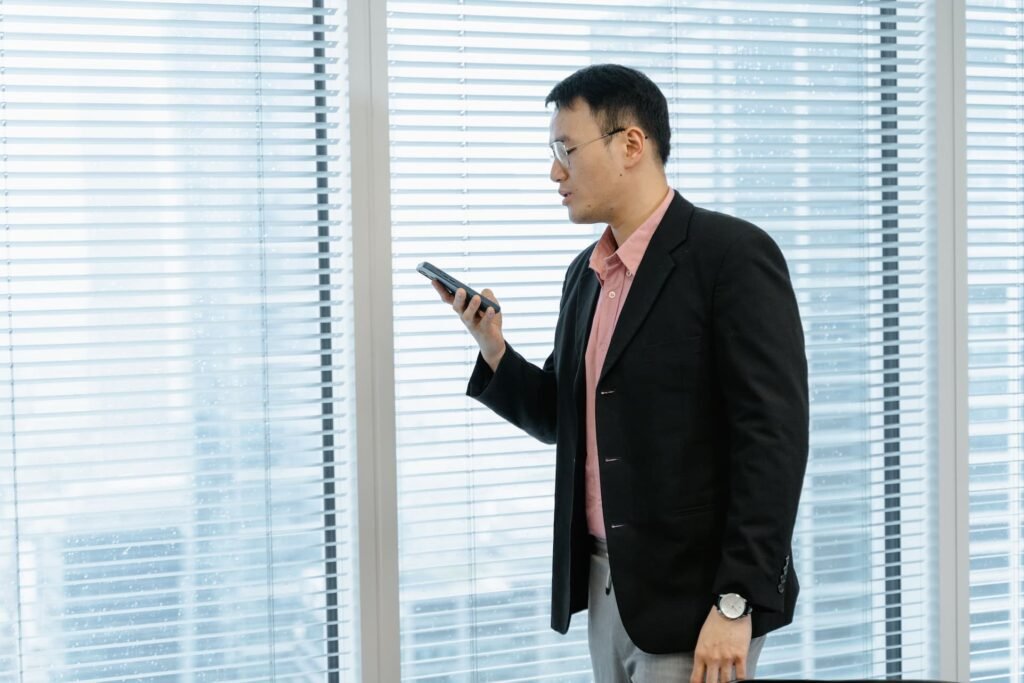 Businessman in a blazer speaking into his phone near an office window, illustrating how B2B buyers use voice and AI search for B2B vendor research