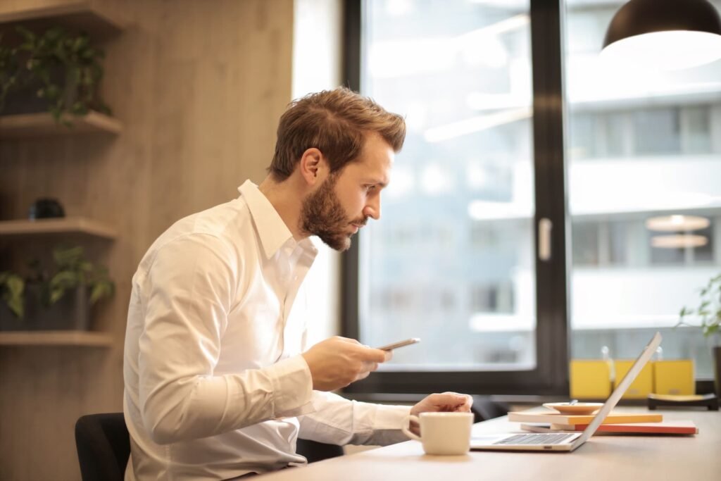Professional at his desk checking his phone beside an open laptop, illustrating how AI search visibility determines which vendors B2B buyers find first